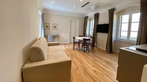 Un salon comprenant un canapé, une table à manger avec des chaises, un téléviseur fixé au mur et une fenêtre avec des rideaux.