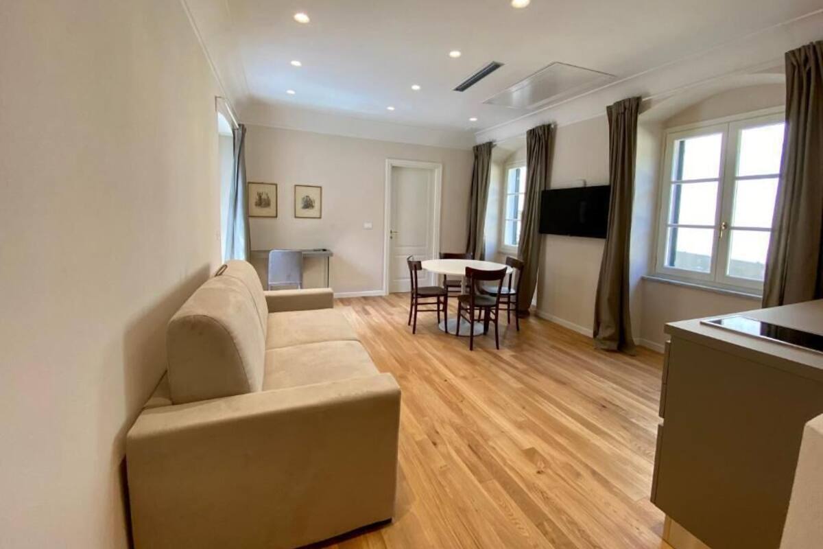 Ein Wohnzimmer mit Sofa, einem Esstisch mit Stühlen, einem an der Wand montierten Fernseher und einem Fenster mit Vorhängen.
