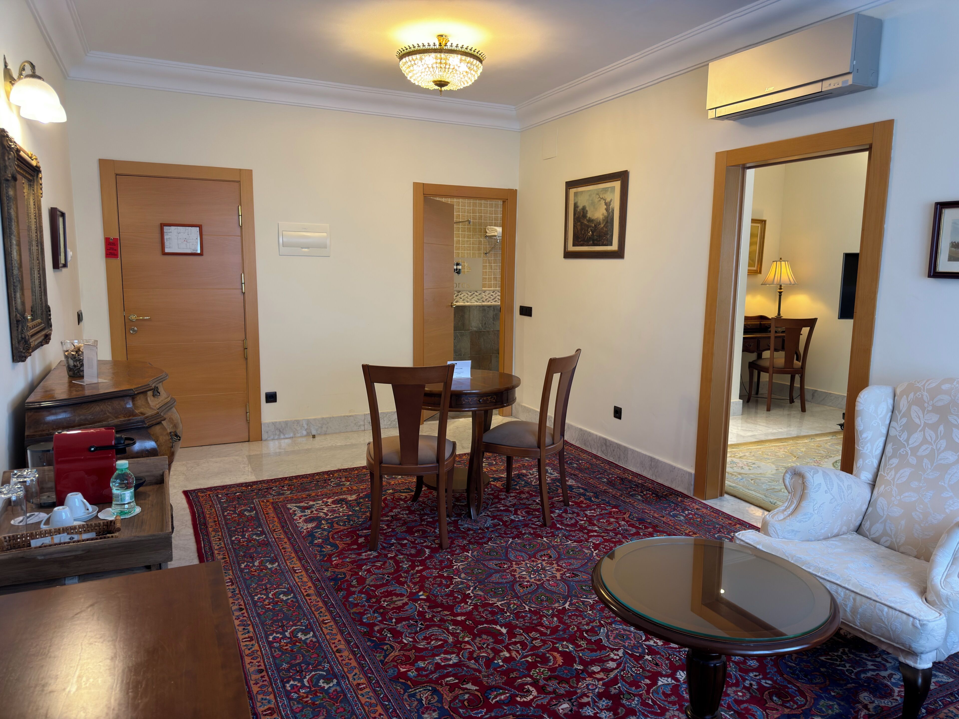 A room with a round glass table, a sofa, a wooden cabinet, a red rug, and a door leading to another room.