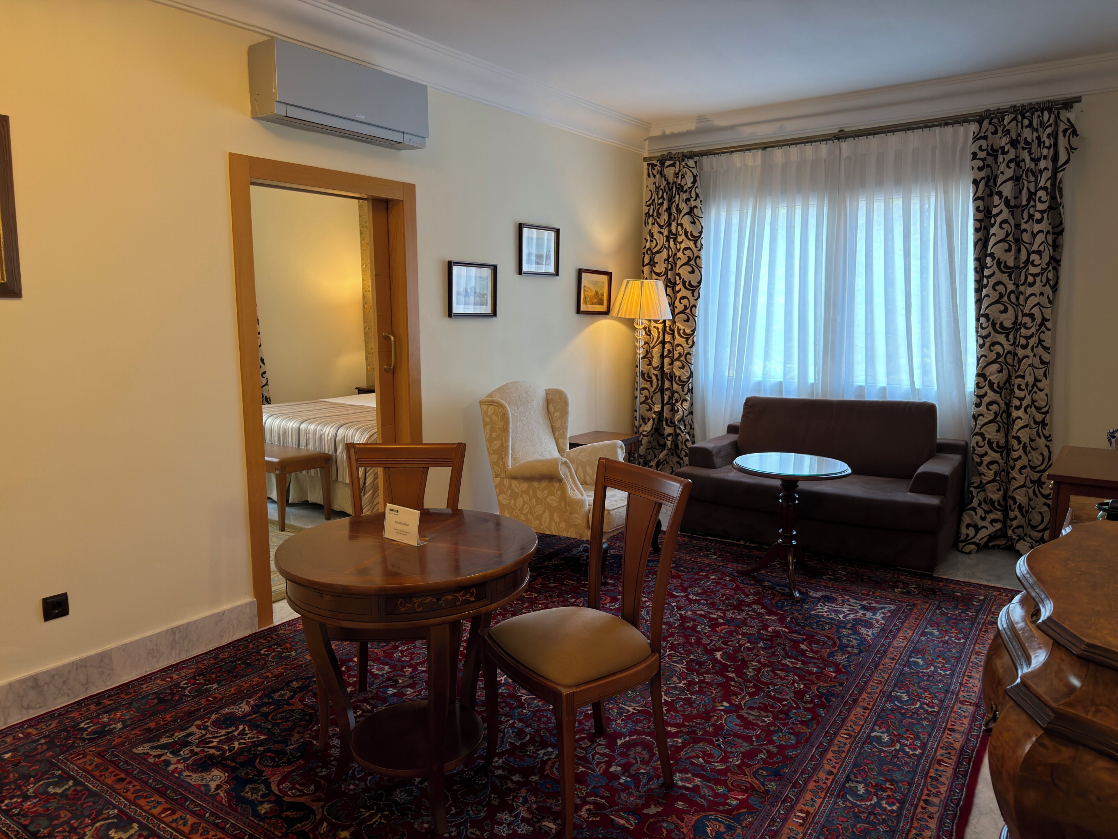 A hotel room with a bed, a sofa, a round table with chairs, a lamp, and a patterned rug.