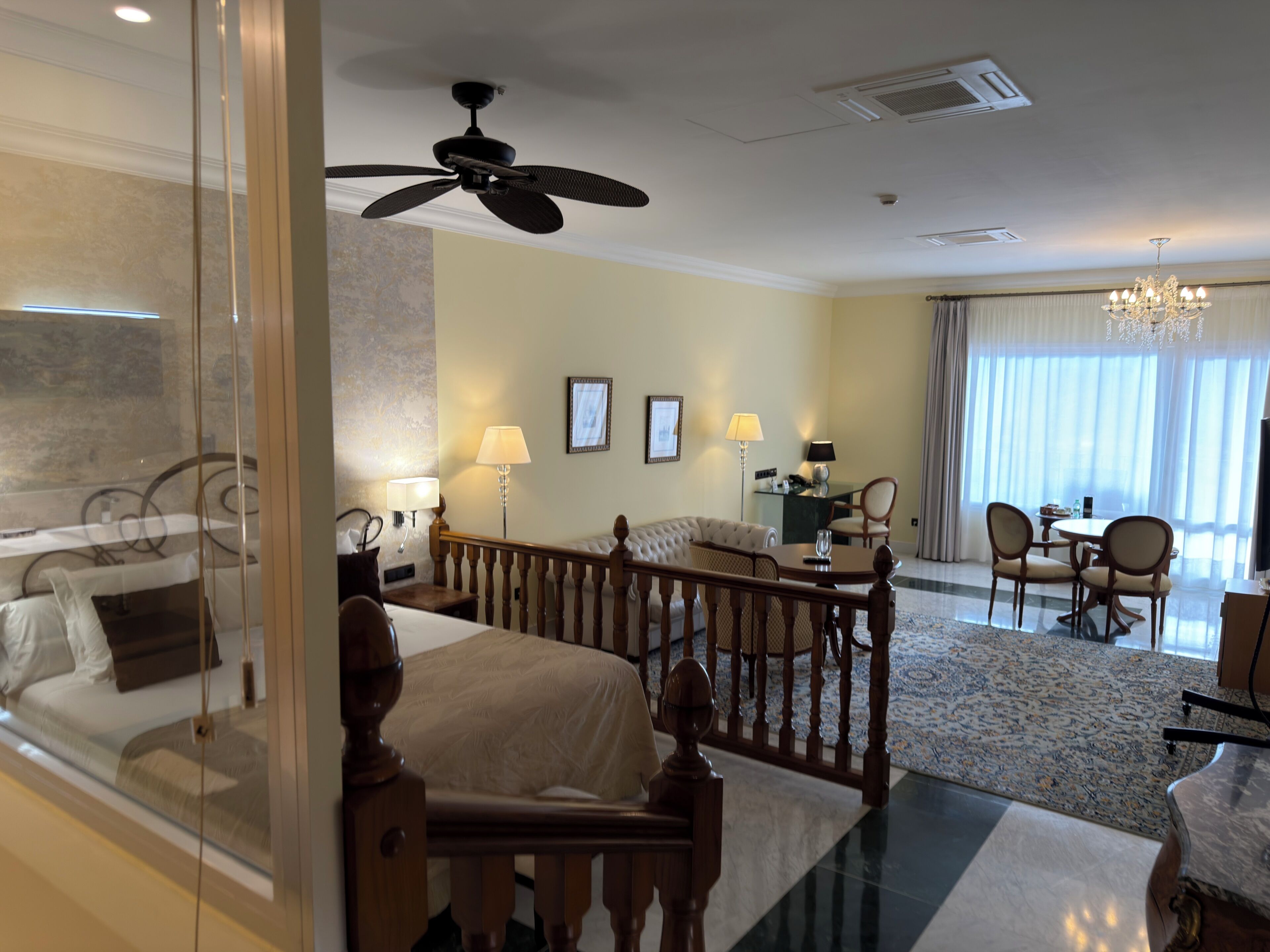 A hotel room with a bed, a desk, a chair, a sofa, a chandelier, and a view of the outdoors.
