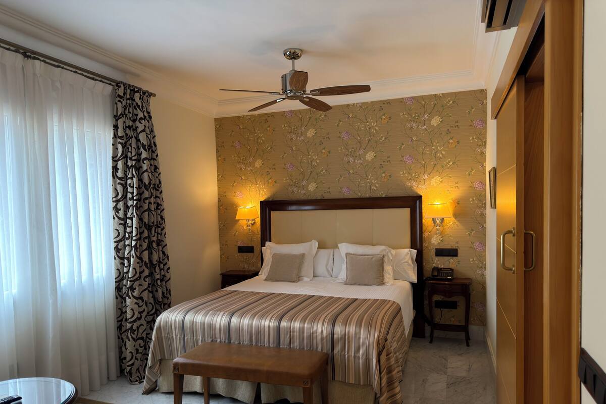 A hotel room with a bed, a ceiling fan, a bedside table with a lamp, a bench, and a round table with a remote control.