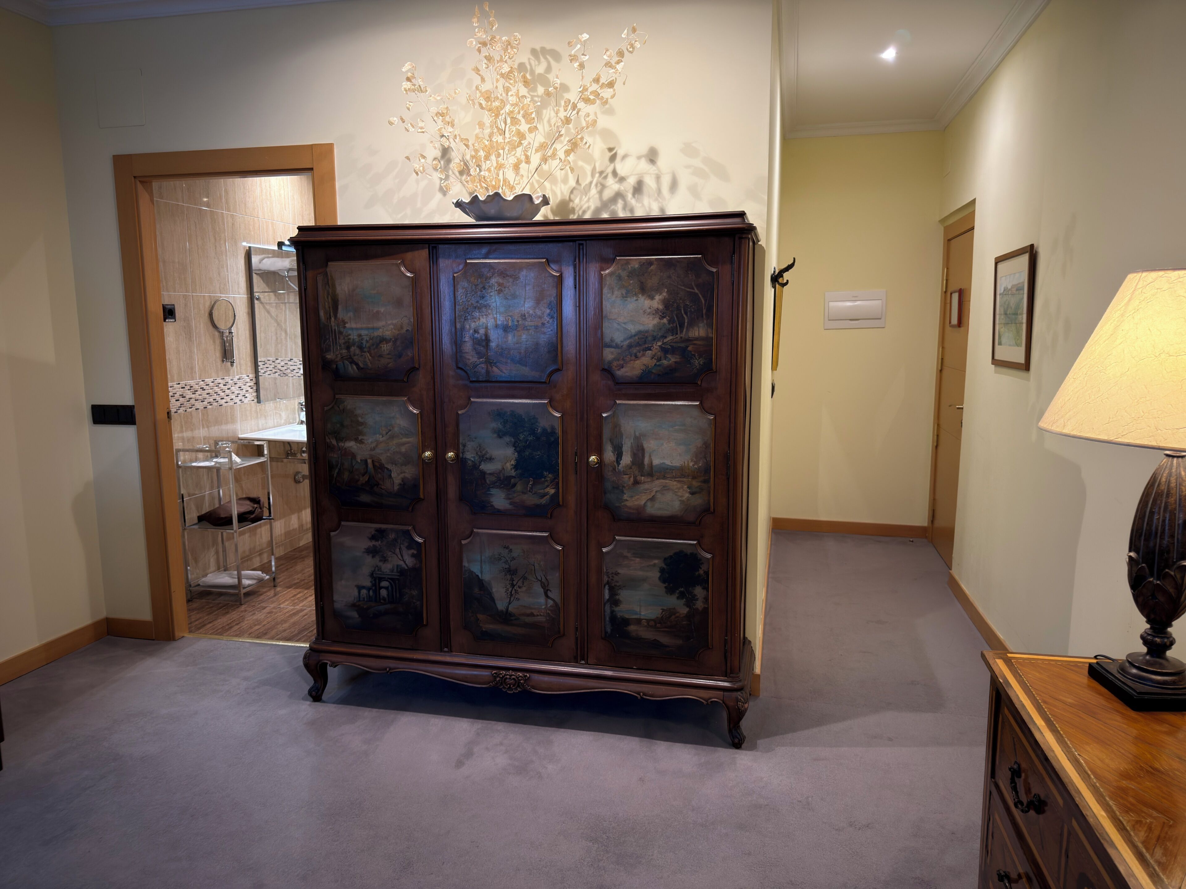 A hallway with a large wooden cabinet featuring glass doors with landscape paintings, a lamp, and a door leading to a bathroom.