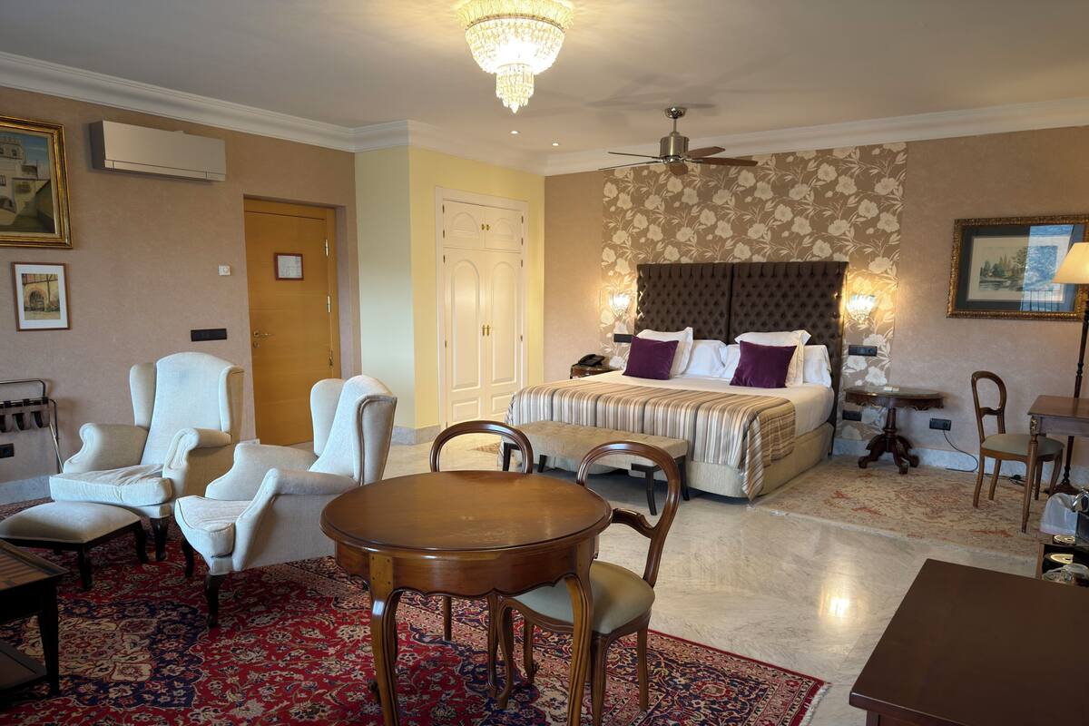 A hotel room with a bed, two armchairs, a wooden table, a chandelier, a ceiling fan, and a patterned carpet.