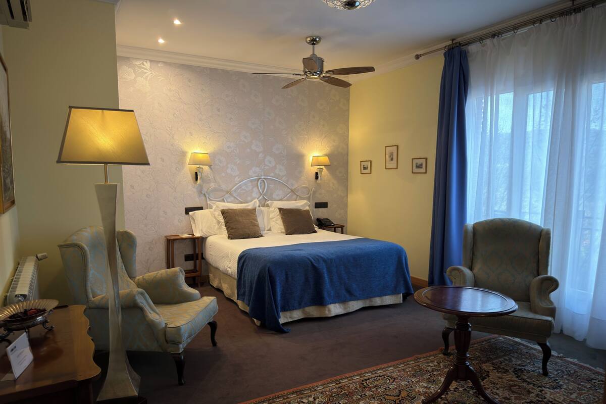 A hotel room with a bed, two bedside lamps, a ceiling fan, a desk with a lamp, two armchairs, and a patterned rug.