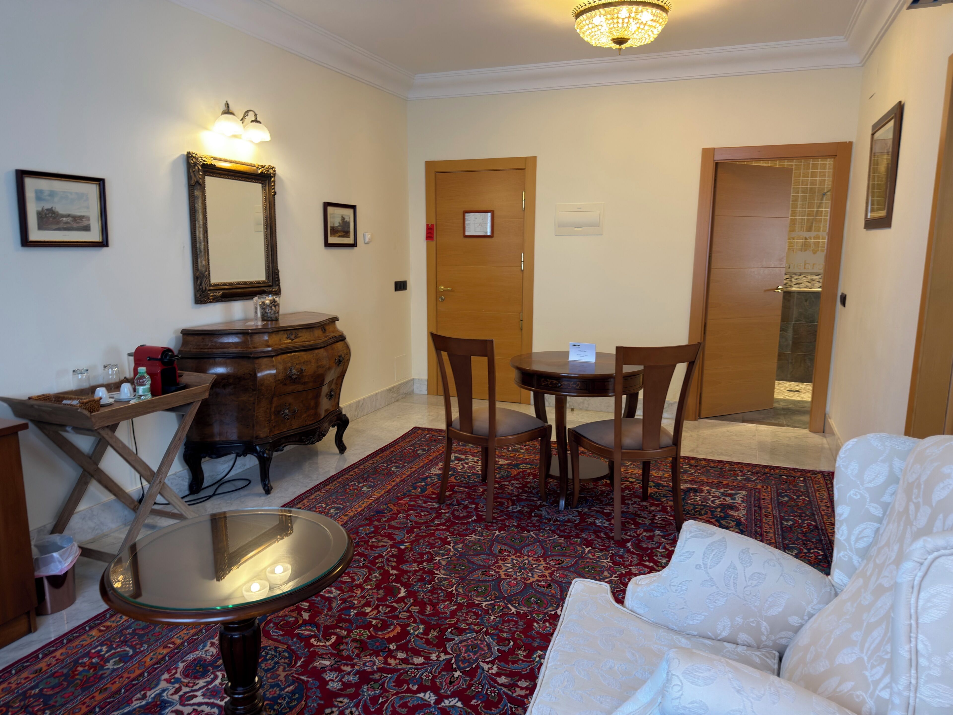 A hotel room with a round table, two chairs, a sofa, a wooden dresser, a mirror, and a red patterned rug.