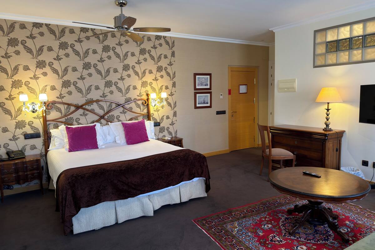 A hotel room with a bed, a desk, a chair, a ceiling fan, a wall-mounted TV, and a patterned rug.