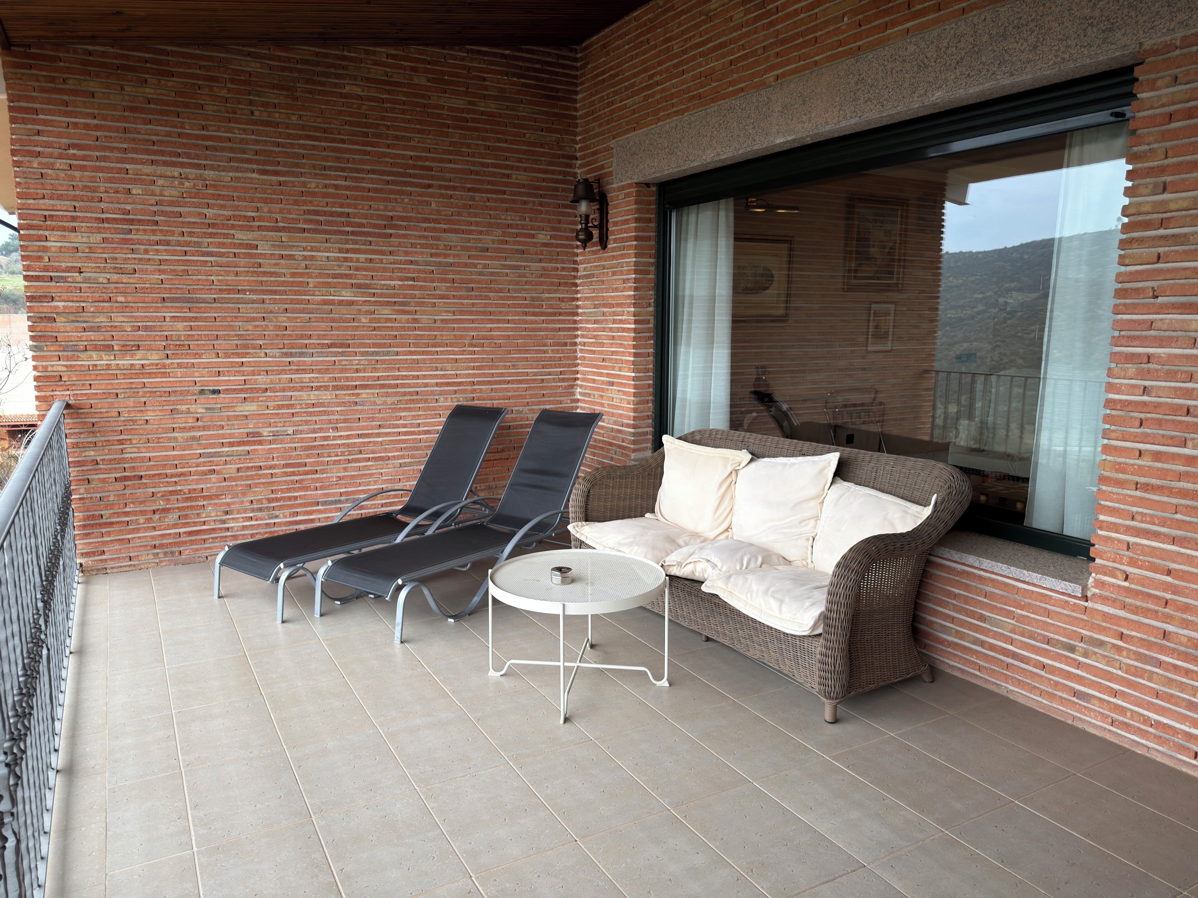 Ein Balkon mit einer Rattan-Couch, zwei Loungestühlen, einem kleinen runden Tisch und Blick auf eine Berglandschaft durch ein großes Fenster.