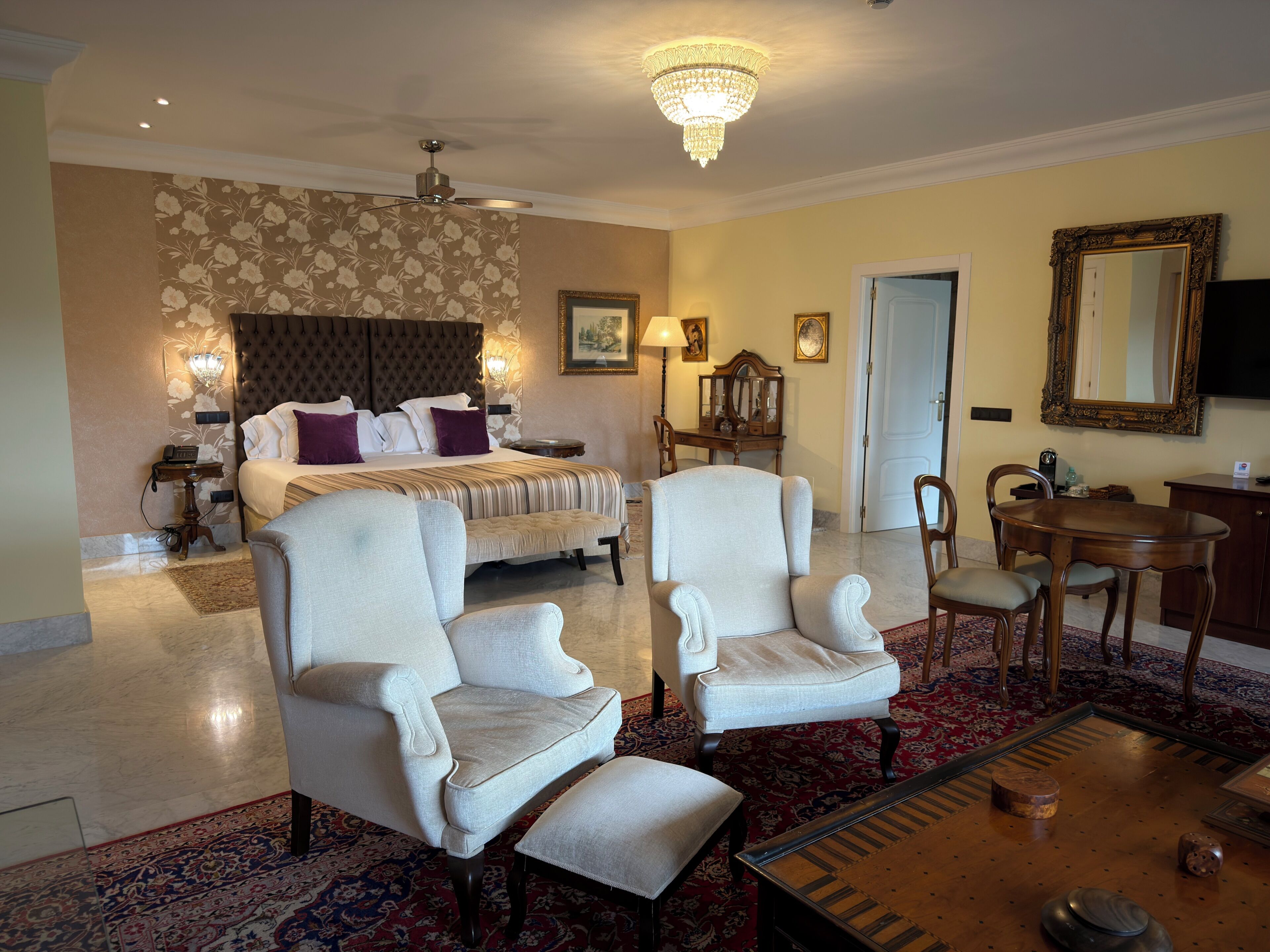 A hotel room with a bed, two armchairs, a desk, a chandelier, and a large mirror.