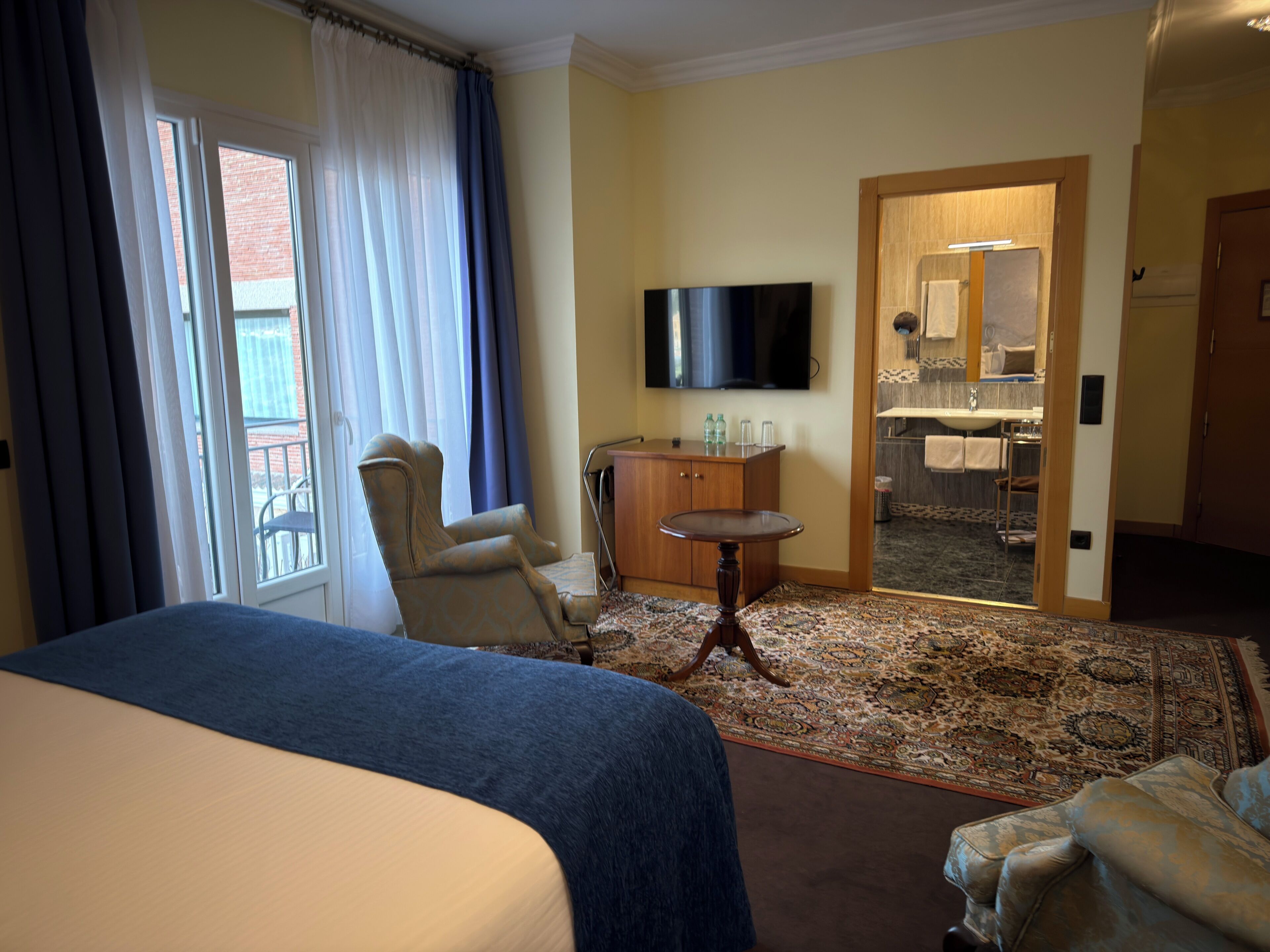 Habitación de hotel con cama, una silla, una mesita, televisor y baño visible a través de una puerta abierta.