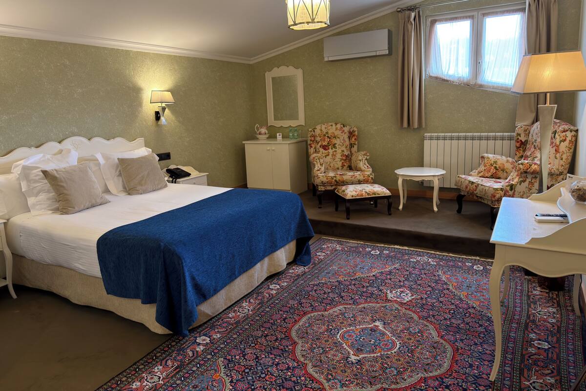 A hotel room with a bed, a desk, a chair, a lamp, a mirror, a radiator, and a window with curtains.