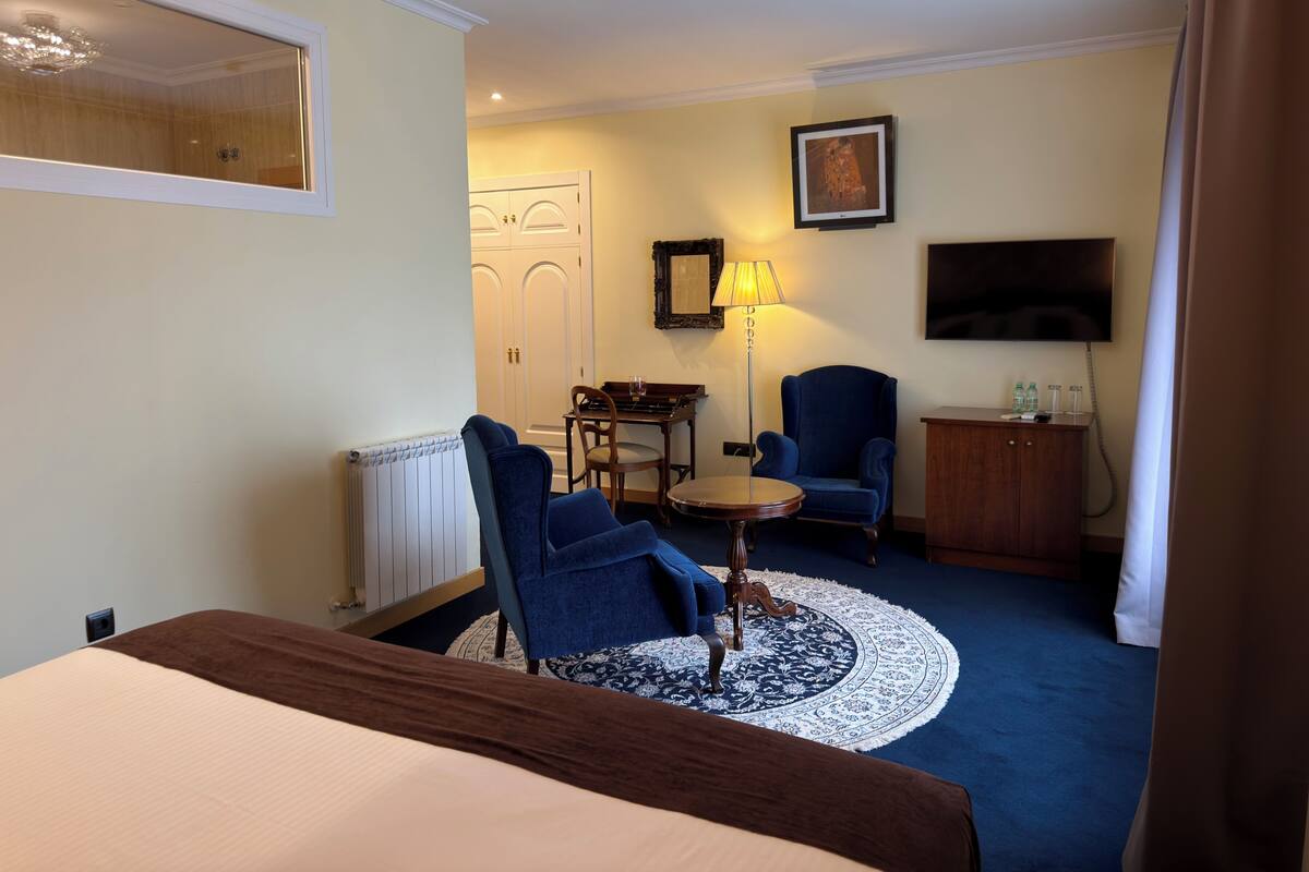 A hotel room with a bed, two blue armchairs, a small round table, a lamp, a television, and a cabinet.
