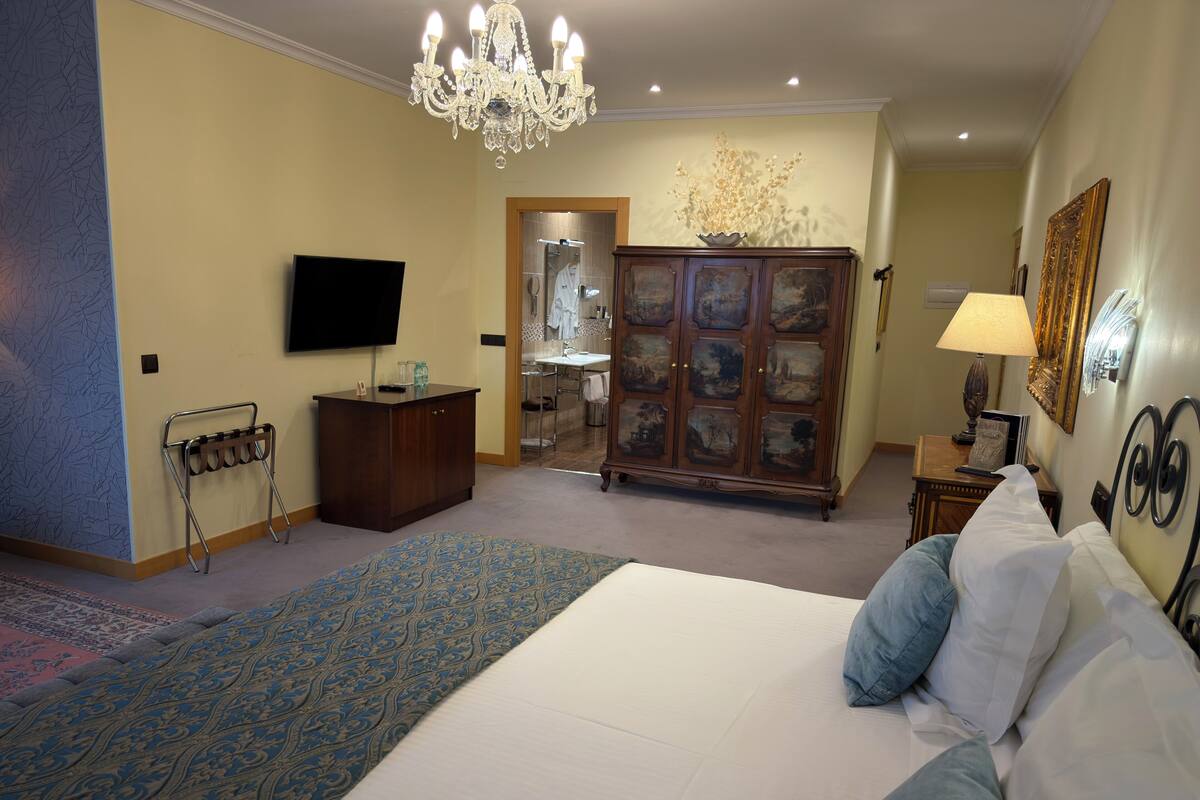A hotel room with a bed, a television, a chandelier, a cabinet with paintings, and a bathroom visible through an open door.