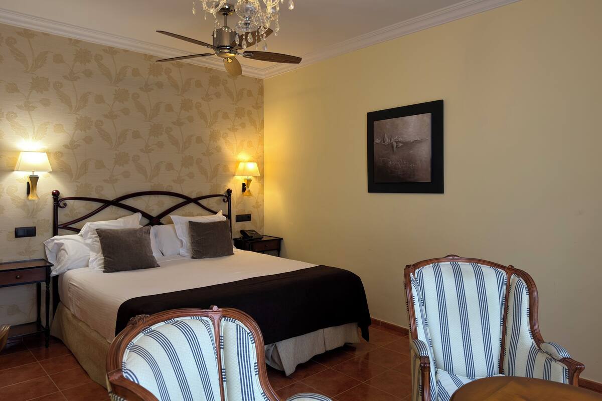 A hotel room with a bed, two bedside tables, a chandelier, a wall-mounted lamp, a framed picture, and two upholstered chairs.