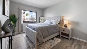 A bedroom with a bed, bedside tables, a window, a plant, and a view of palm trees. - Meticulously Kept Downtown Mesa 1-Bedroom Oasis (Mesa)