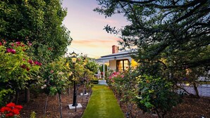 A garden pathway leading to a house with a white facade and a red roof, surrounded by lush greenery and flowering plants. - Luxury Glam Pool Retreat Near Glenelg Beach-sleeps 10 (Dover Gardens)