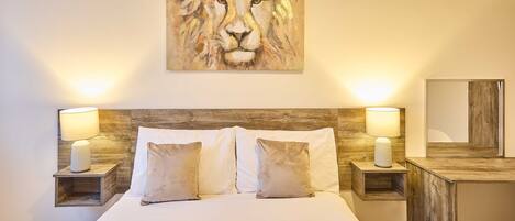 A bedroom with a large bed, two bedside lamps, a wooden headboard, a painting above the bed, and a mirror on the wall.
