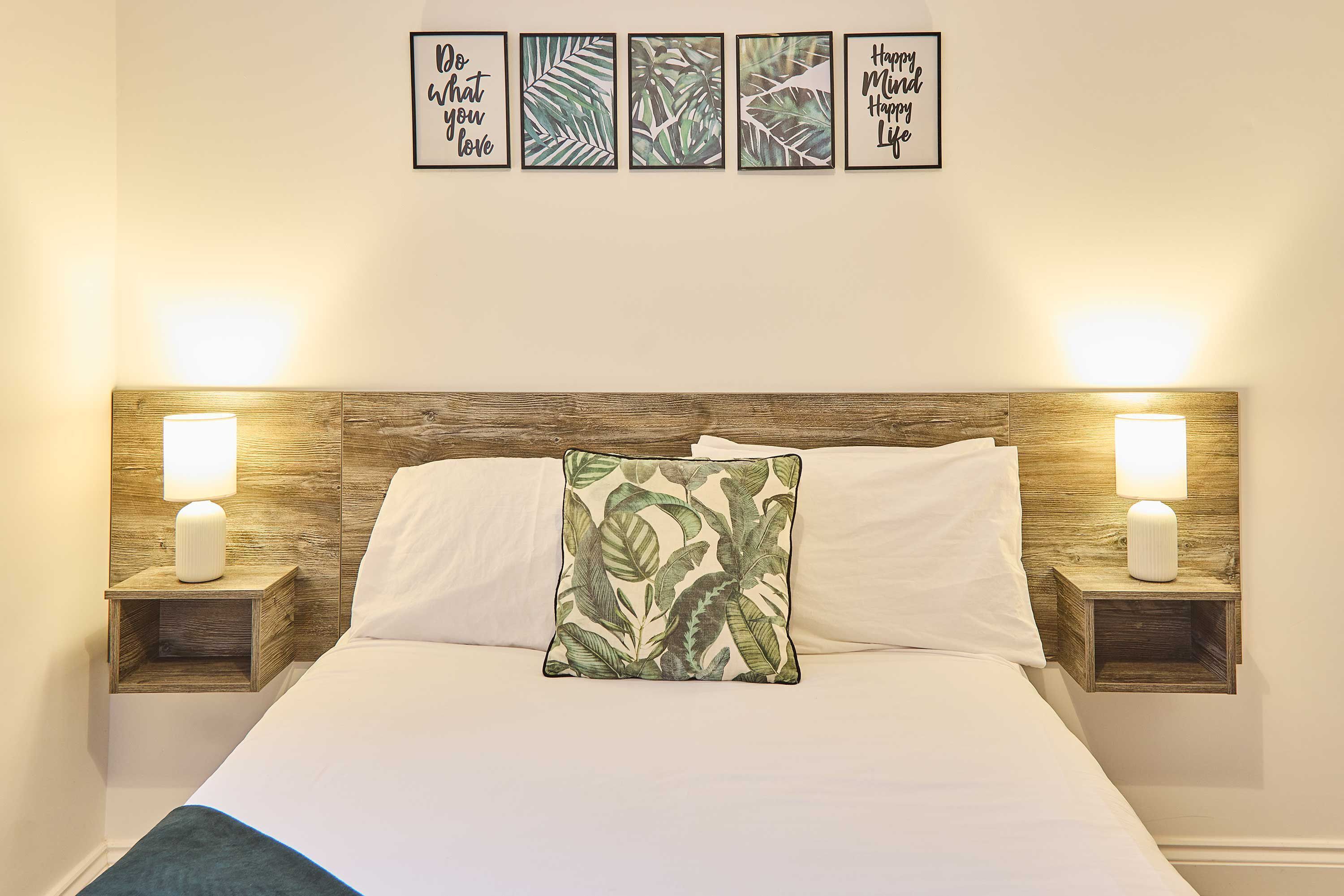 A neatly made bed with a white comforter, two white pillows, and a green leaf-patterned pillow. There are two bedside tables with white lamps, and a wall with four framed motivational quotes.