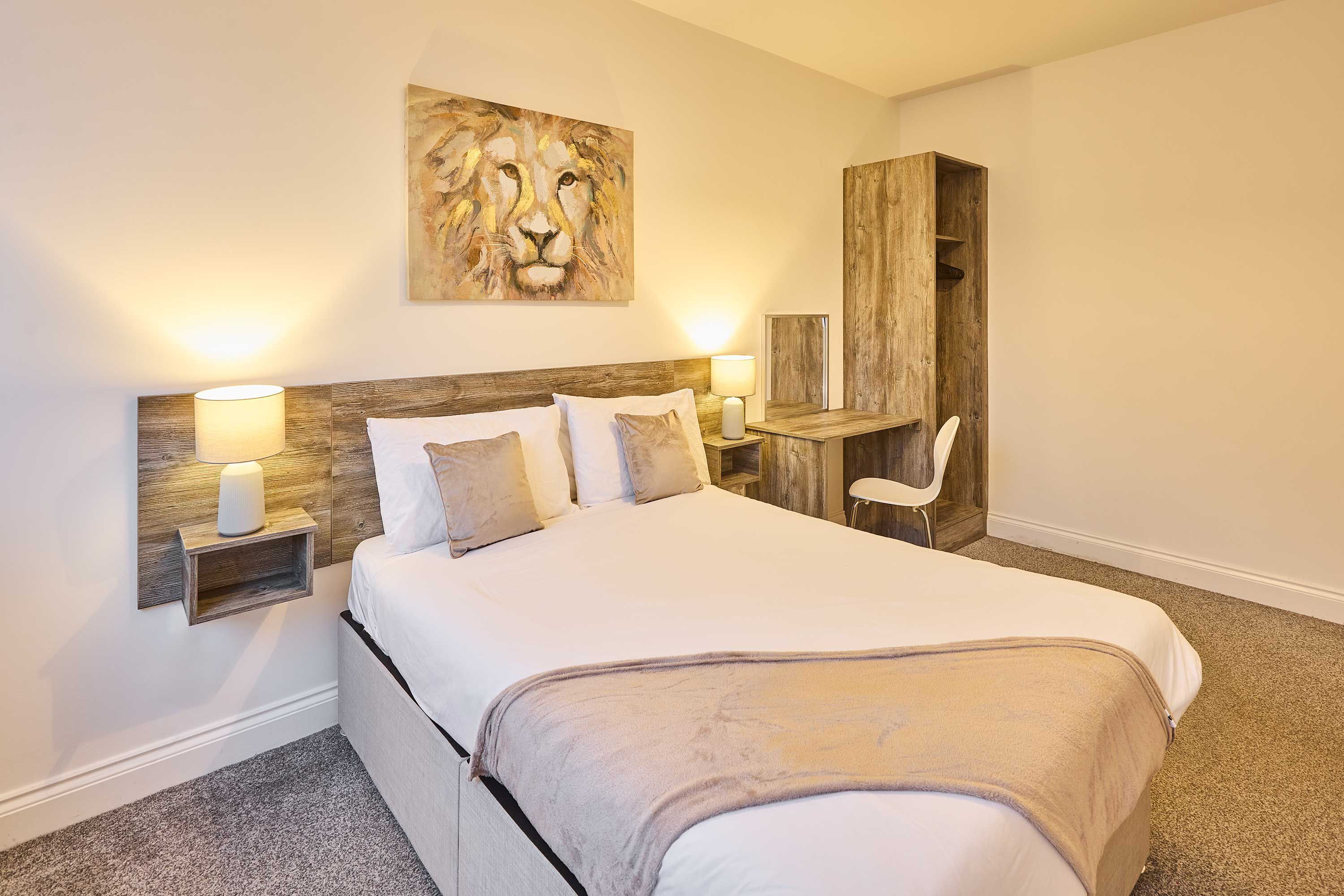 Une chambre avec un grand lit, des lampes de chevet, une tête de lit en bois, un bureau avec une chaise et un tableau représentant un lion accroché au mur.