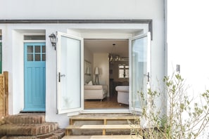 Um edifício branco com porta azul, escadaria de madeira e um quarto com cama e abajur.