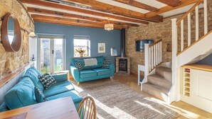 Un salon avec un mur en pierre, des poutres apparentes, un canapé bleu, une cheminée et un escalier.