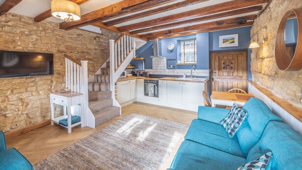 Un intérieur rustique avec des poutres apparentes, un mur en pierre, un canapé bleu, une cuisine et un escalier.