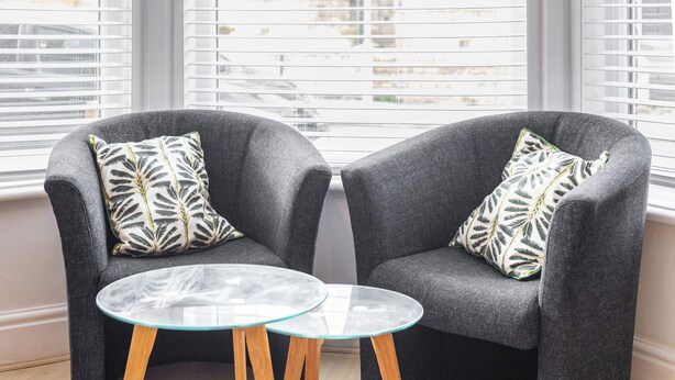 Two grey armchairs with leaf-patterned cushions, a glass table with wooden legs, and a window with blinds. - Host Stay Elmville (Scarborough)