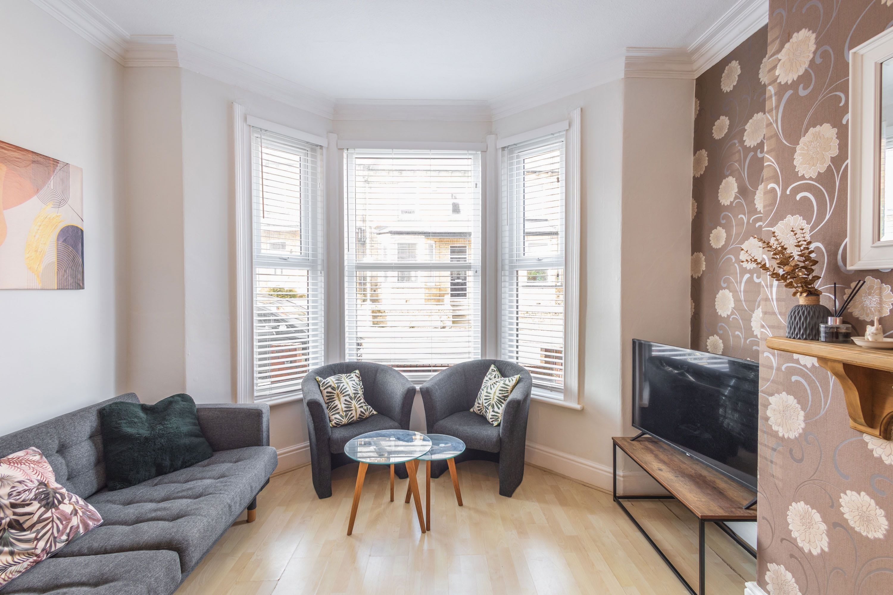 A living room with a sofa, two armchairs, a glass coffee table, a TV, and a bay window.