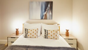 Een bed met witte lakens, gekleurde kussens, een grijze plaid en een schilderij erboven.