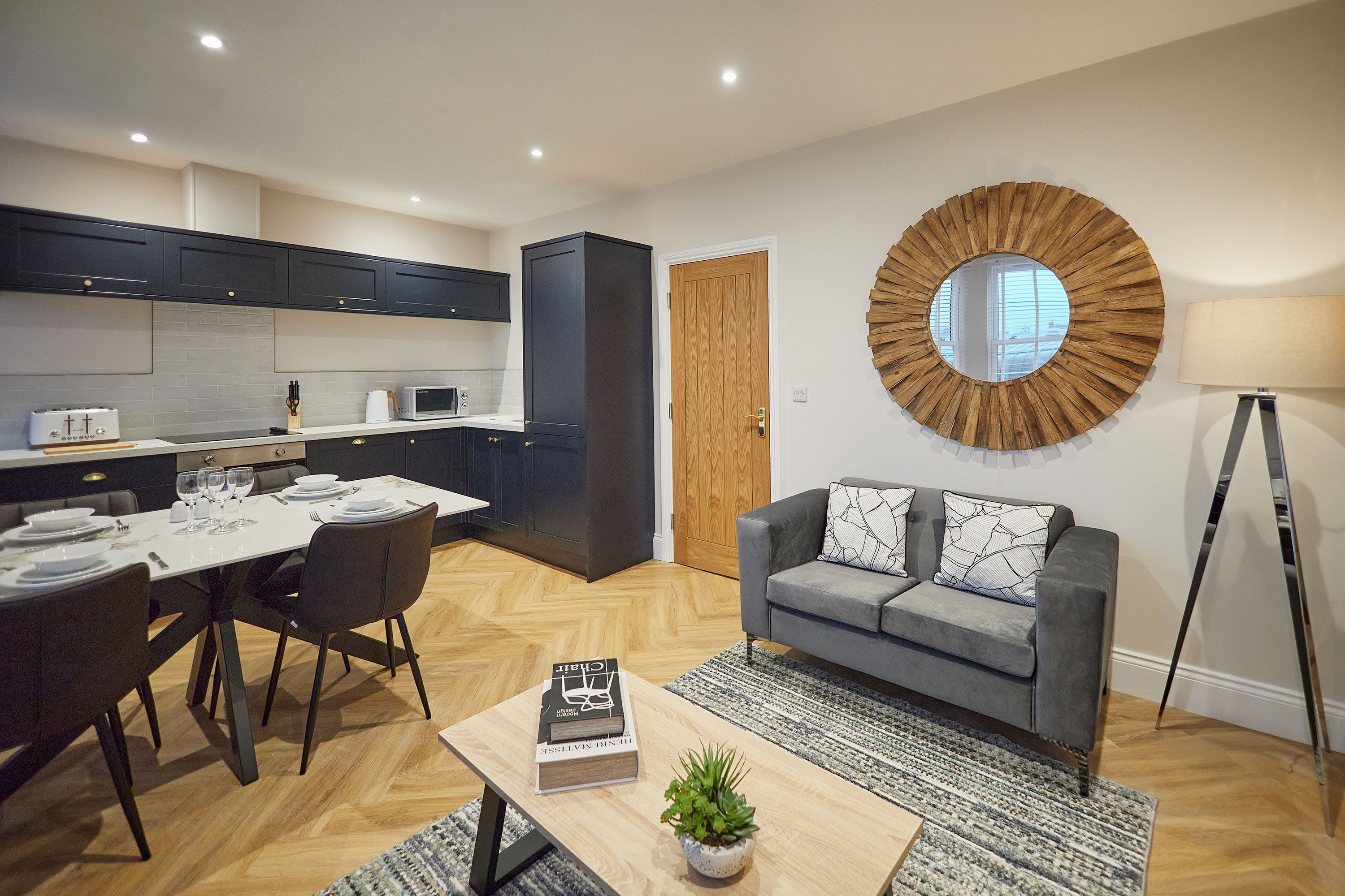 Une cuisine moderne avec des meubles de couleur sombre, un coin repas avec une table blanche dressée, un canapé gris et un miroir rond accroché au mur.