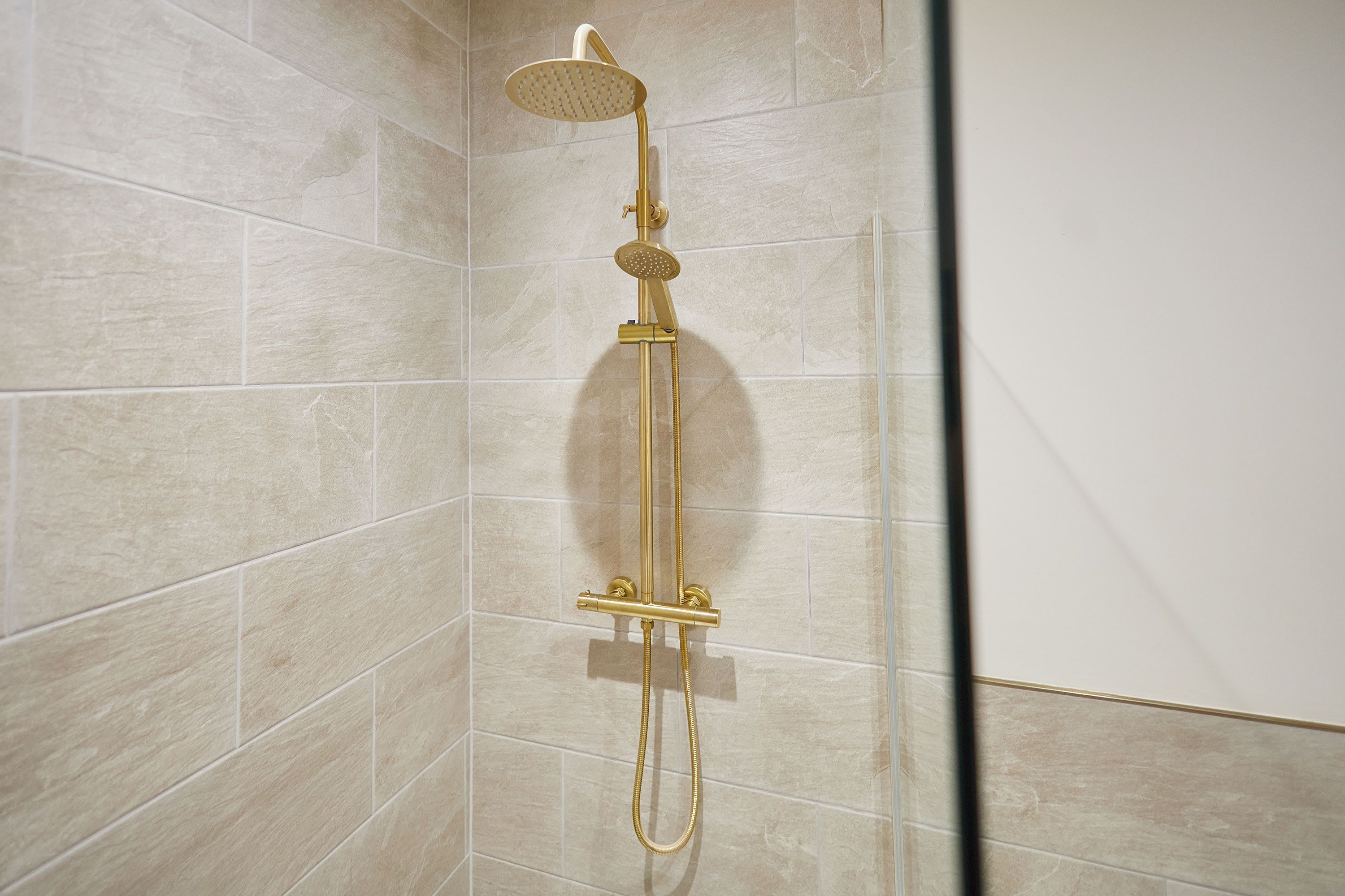 Ducha con cabezal de ducha y manguera de mano, fijada en una pared con azulejos color beige.