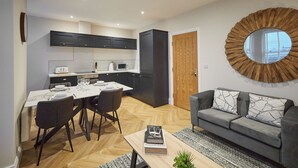 Een moderne keuken met donkere kastjes, een eethoek met een witte tafel en stoelen, een grijze bank met gekleurde kussens, en een ronde spiegel aan de muur.