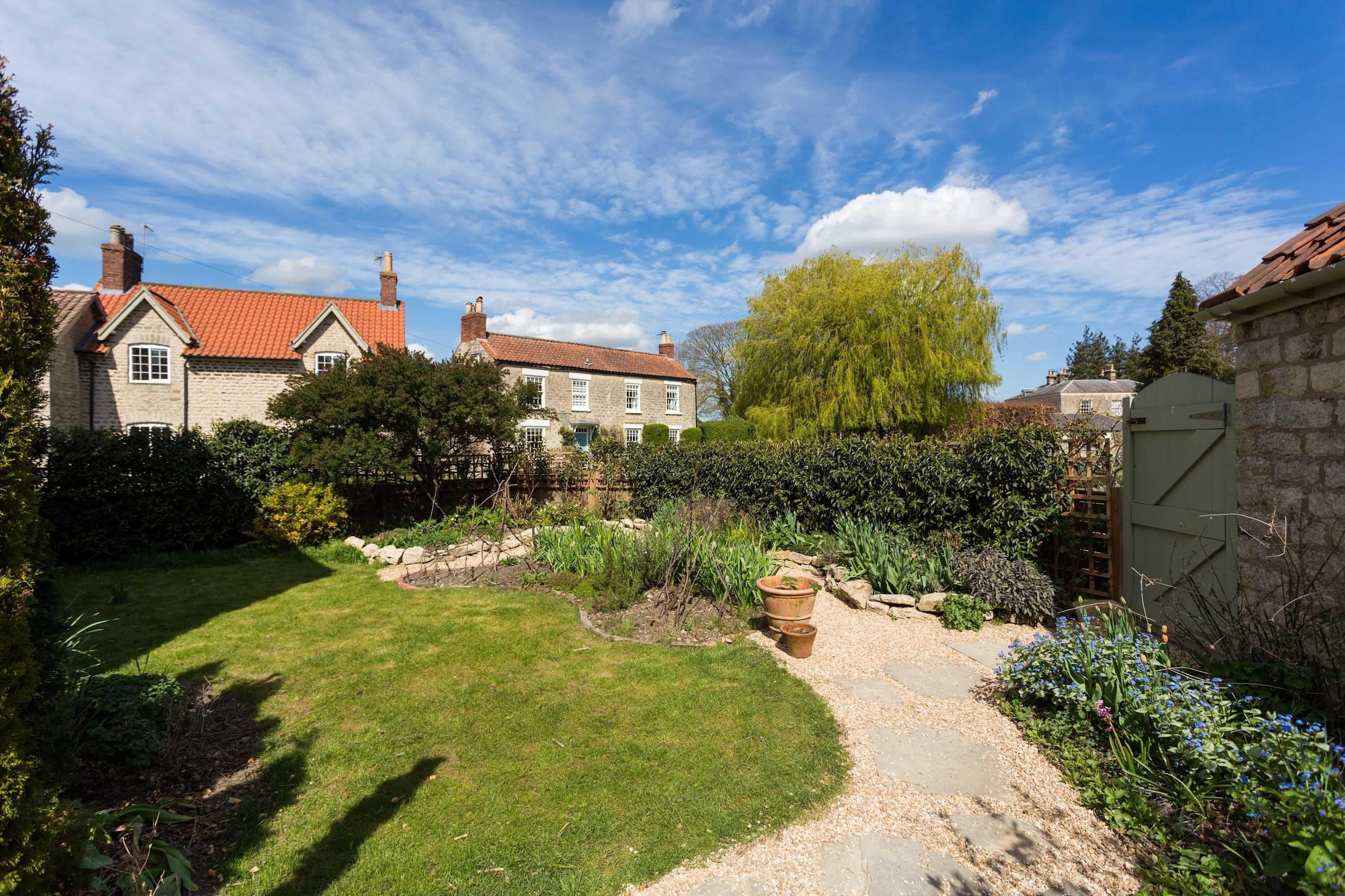 A stone house with a red-tiled roof, a garden with a gravel path, and a large tree.