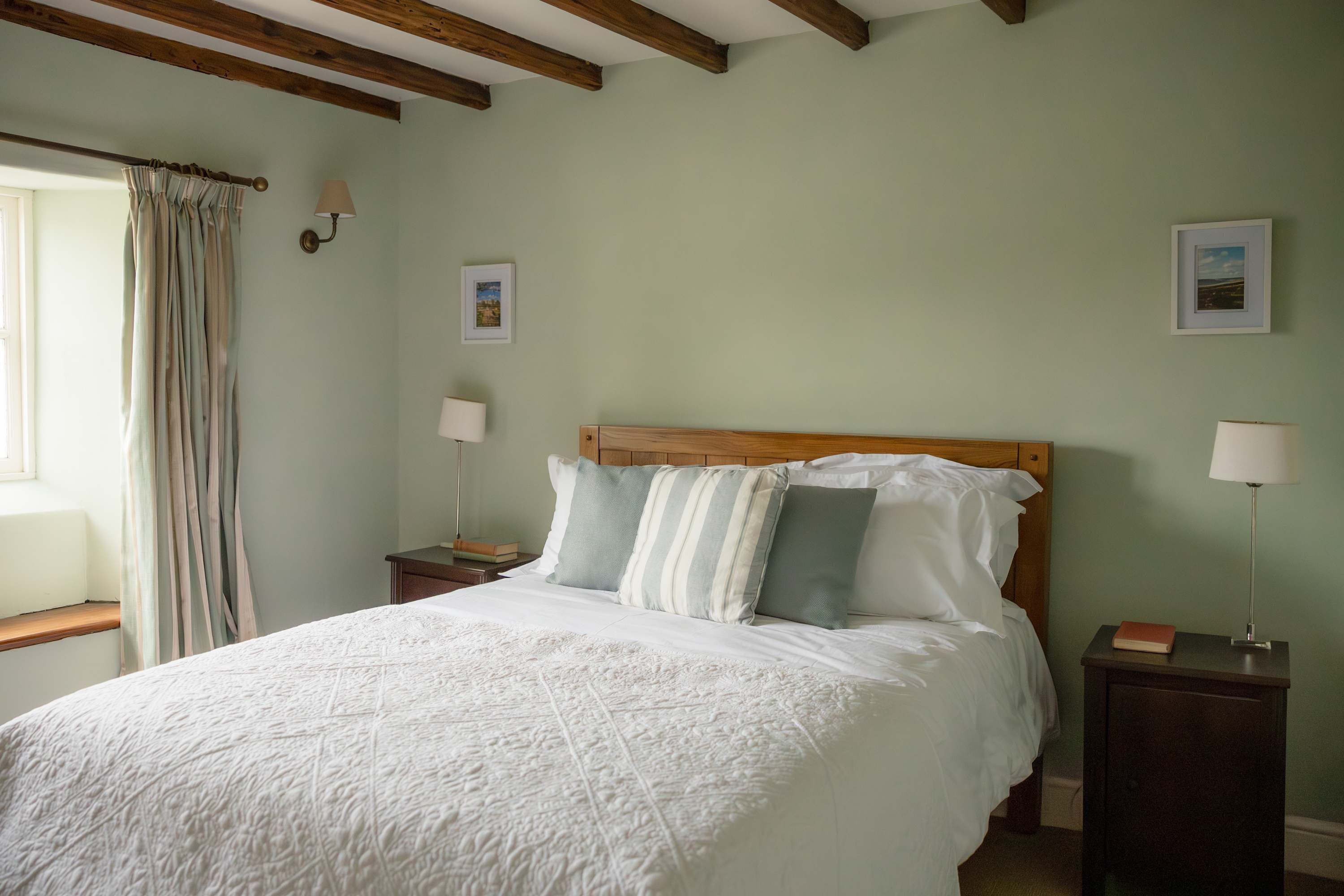 A bedroom with a bed, two bedside tables, a window with curtains, and a wooden ceiling beam.