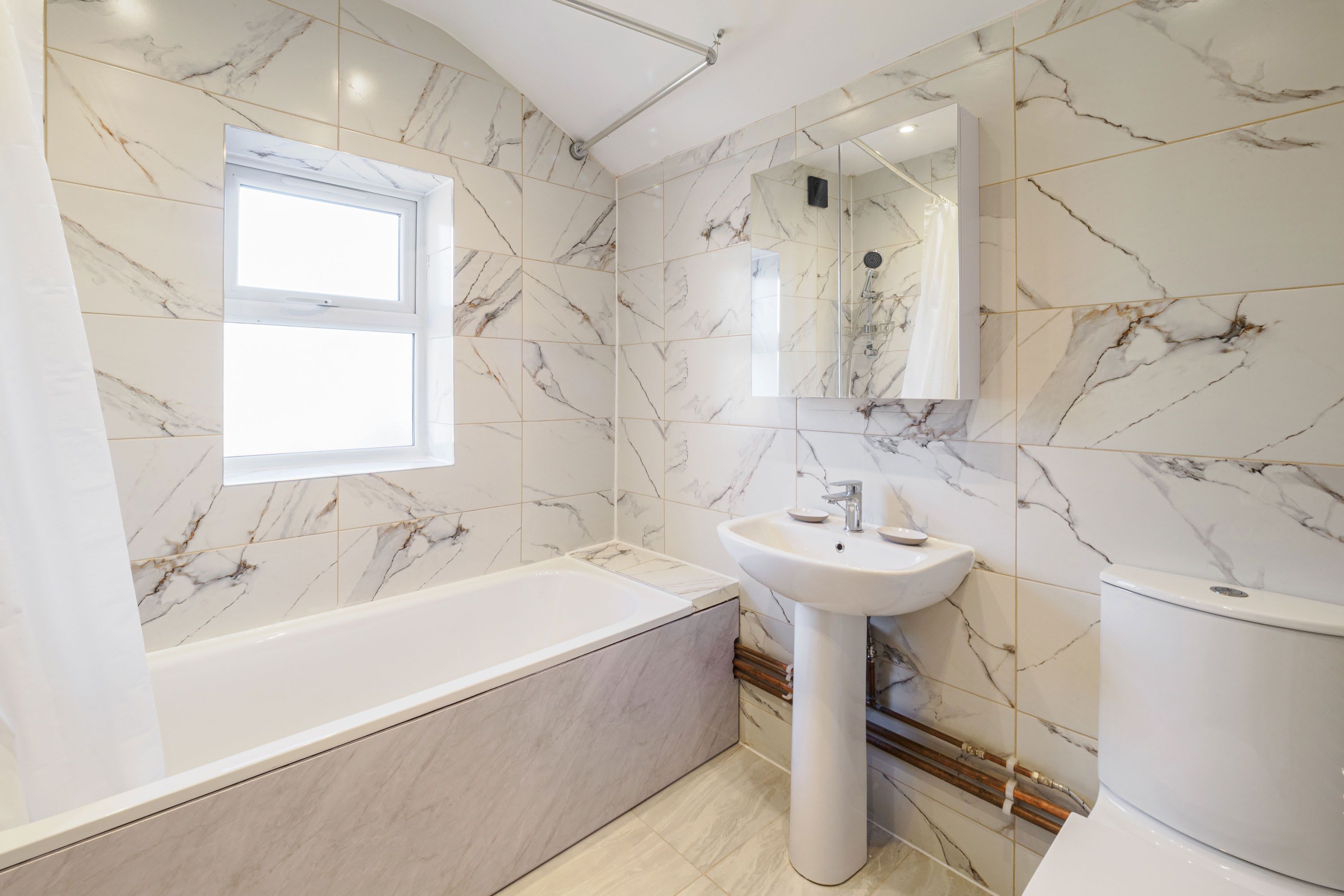 A modern bathroom with a bathtub, sink, and toilet, featuring marble walls and a window.