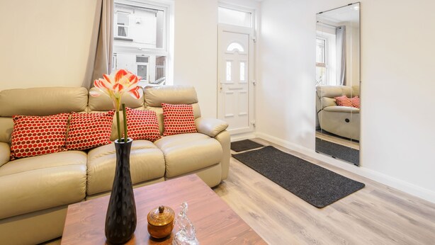 A living room with a beige sofa, red and white patterned pillows, a vase with flowers, a wooden coffee table, a mirror, and a view of a building through a window. - Host Stay Roach Street (Rochester)