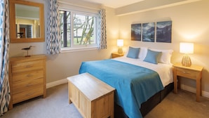A bedroom with a bed, bedside tables, a dresser, a bench, a window with curtains, and wall art. - Host Stay 11 Troutbeck (Penrith)