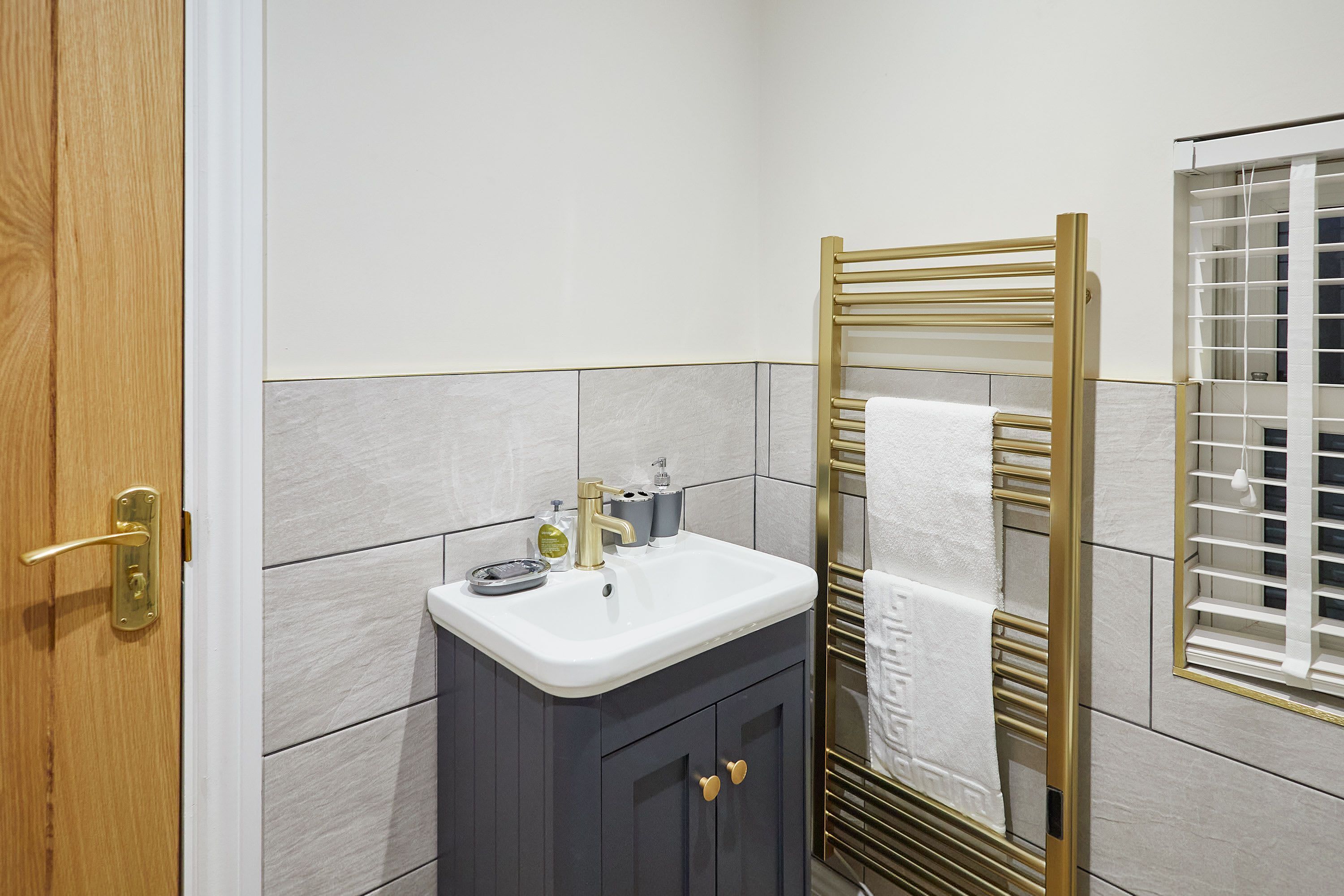 A bathroom with a white sink, dark cabinet, gold towel rack, and a window with blinds.