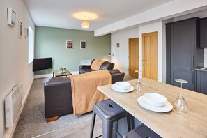Ein modernes Wohnzimmer mit einer Couch, einem Esstisch mit Tellern und einem Weinglas, einem Fernseher und einer Küchenzeile mit Schränken.