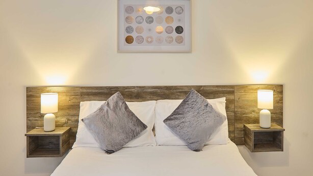 Cama bem arrumada com duas almofadas, uma obra de arte emoldurada acima e duas luminárias de cabeceira em mesas de cabeceira de madeira.
