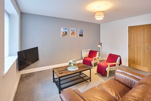 Ein Wohnzimmer mit Fernseher, Couchtisch mit Vase, zwei rote Sessel, ein braunes Ledersofa und eine Holztür.