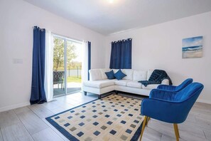 Ein modernes Wohnzimmer mit einem weißen Sofa, blauen Sesseln, einem gemusterten Teppich und einer Glastür, die nach draußen führt.