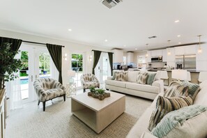 Een moderne woonkamer met een bank, fauteuils, een salontafel en een keukenhoek met kasten en apparatuur.