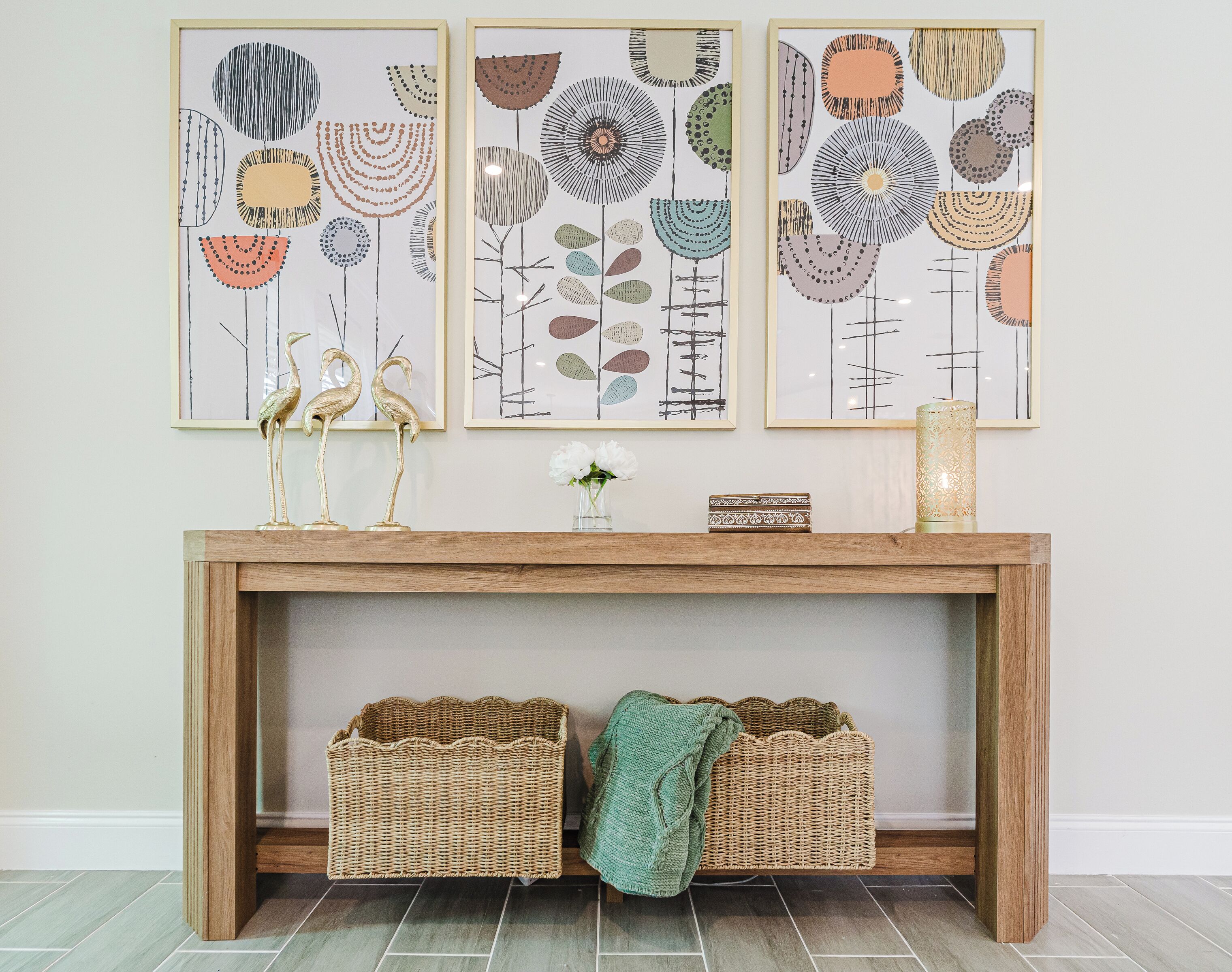 A wooden console table with three framed abstract paintings, two wicker baskets, a vase with flowers, and a decorative lamp.