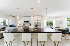 A modern kitchen with white cabinets, a large island with bar stools, stainless steel appliances, and pendant lighting. - Brand new 3 bedroom home with huge pool and garden (Lake Worth Beach)