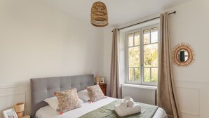 Cama bem arrumada com cabeceira cinza, mesa de cabeceira com abajur, janela com cortinas e espelho fixado na parede.