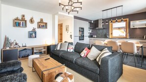 A modern living room with a sofa, coffee table, and a dog. There is a kitchen area with a dining table and chairs. - Les 3 Marmottes - Au pied des Pistes (Huez)