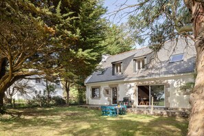 A house with a stone exterior, a covered patio with a table and chairs, and a large tree in the foreground. - Chez Mab - Maison familiale les pieds dans l'eau (Saint-Armel)