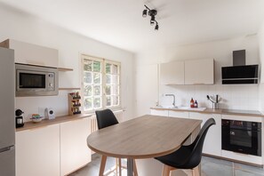 A modern kitchen with a wooden table, black chairs, a microwave, oven, and a window with a view of greenery. - Chez Mab - Maison familiale les pieds dans l'eau (Saint-Armel)