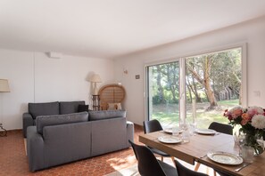 En moderne stue med en grå sofa, et spisebord dækket til to personer, og et stort vindue med udsigt til træer.